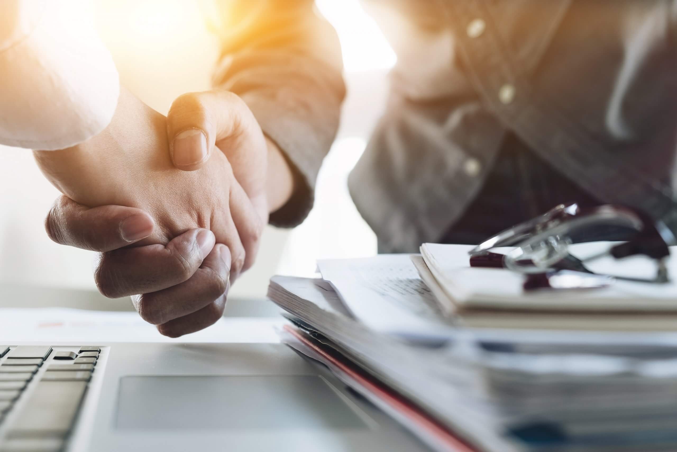Dos profesionales estrechándose las manos en señal de acuerdo sobre un escritorio de oficina con documentos y un portátil